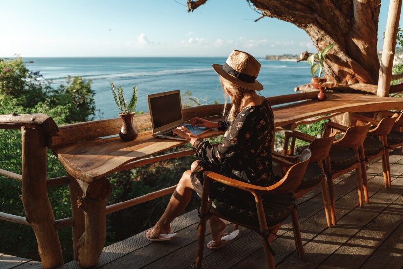 A woman is working on her laptop from a long wood table overlooking a view of the ocean.