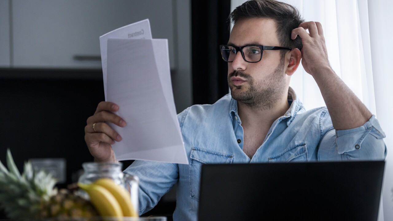 Man looking at paperwork sitting at table with computer, looking confused