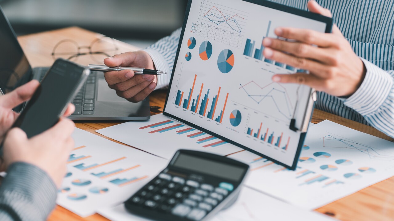 One man presents data charts to another over a desk full of printouts of graphs beside a laptop and a calculator, signifying financial planning