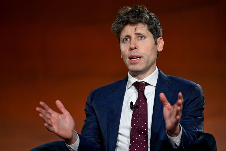 Sam Altman gesturing with open hands while speaking, wearing a blue checkered suit and a maroon polka-dot tie.