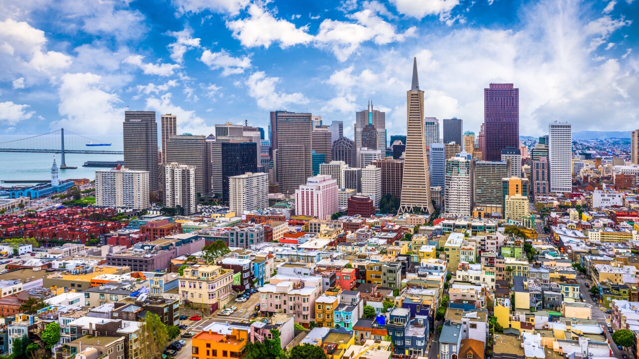 The San Francisco skyline, with multi-colored homes in the foreground before the tall skyscrapers, the sky is blue, and the bay can be seen in the left-hand corner.