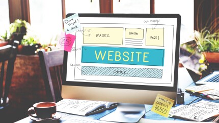 A sketch on a computer monitor surrounded by notebooks, laptops and post-it notes in an office reads "website," symbolizing business website strategy