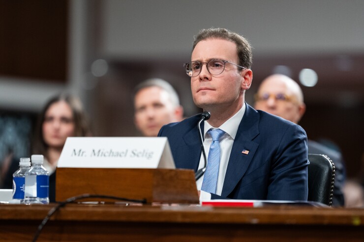 Michael Selig, wearing glasses and a suit, sits at a witness table with a nameplate during a formal hearing or testimony.