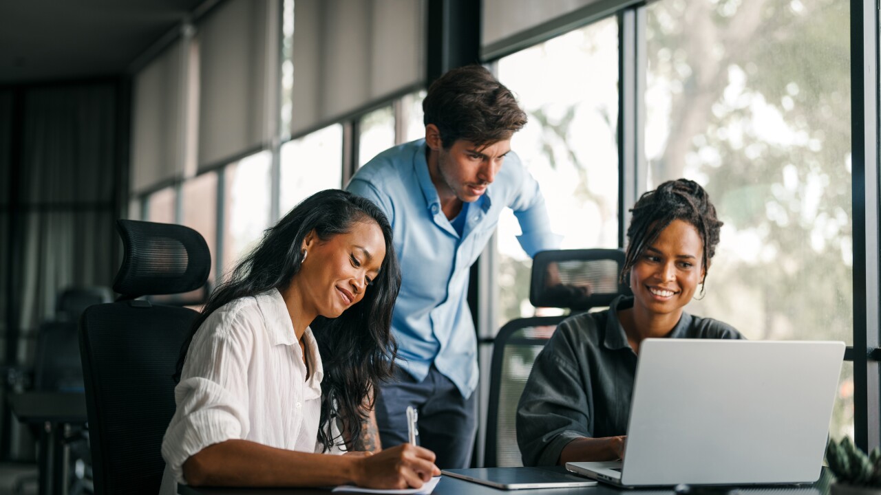 Three employees working together, looking at computer, writing, smiling