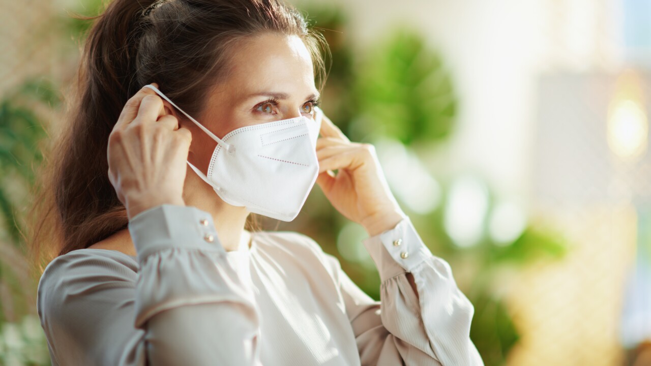 A woman in a grey blouse is putting on a KN95 mask.