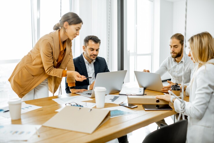Group of men and women at table working with laptops; woman standing with group at work