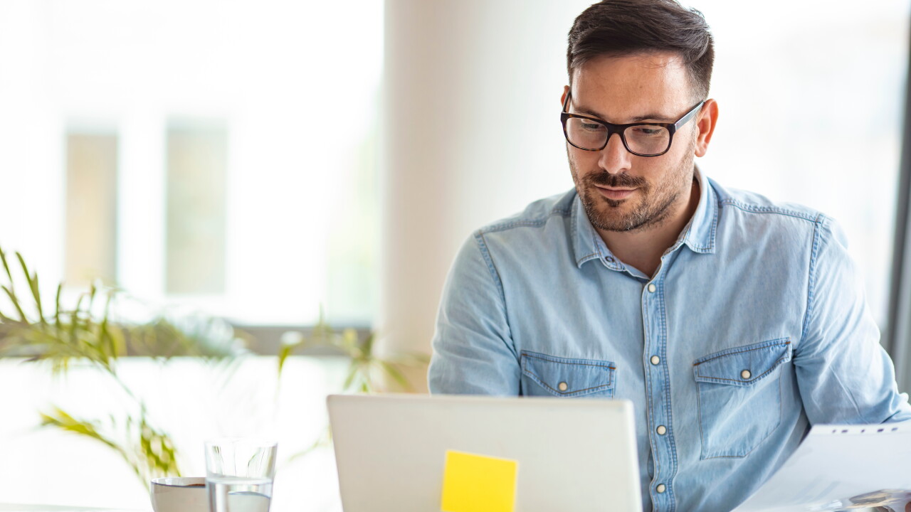 Man with glasses sitting at table desk working on laptop paper in hand