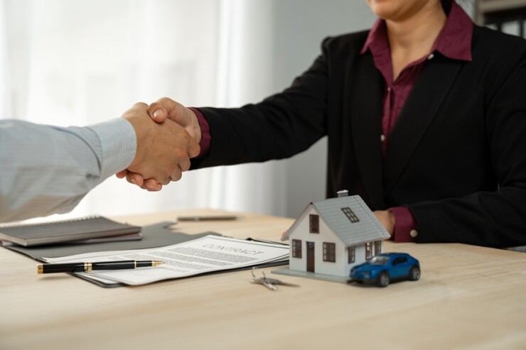 People shaking hands over an insurance policy with a small house and car to the side of the contract.
