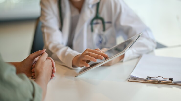 A doctor shows a patient something on an iPad while seated at a table; their heads are not in frame.