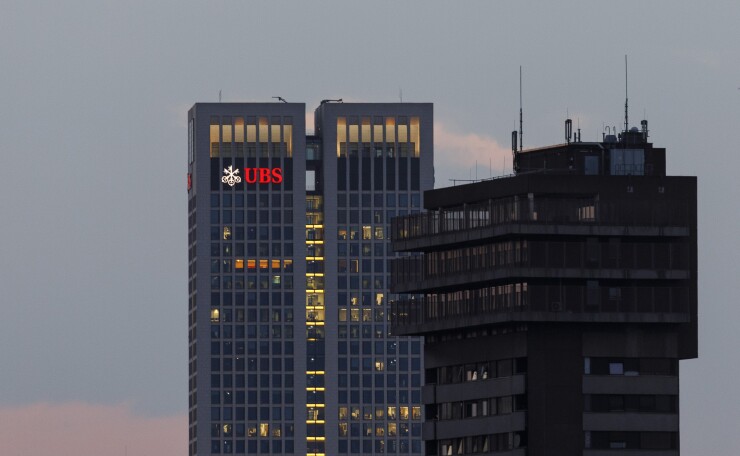 The UBS Group AG logo sits illuminated on the bank's skyscraper offices at dusk in Frankfurt, Germany, on Tuesday, July 17, 2018. Frankfurt's efforts to attract bankers escaping Brexit are in danger of losing momentum. Photographer: Alex Kraus/Bloomberg