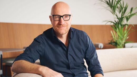 Bald man wearing black glasses and a blue collared shirt sits on beige couch with tall green plant in background.