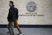 A picture of two people walking past signage outside the Office of the Comptroller of the Currency headquarters in Washington, DC.