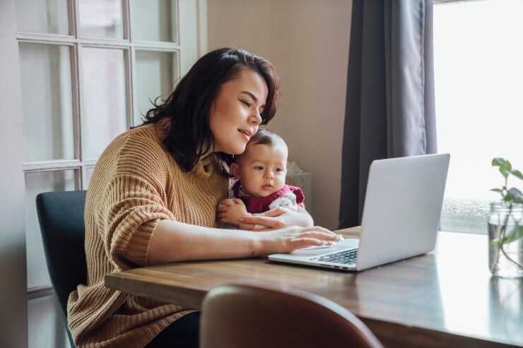 A mom, Latina, sits at her dining room table with her baby boy while scrolling on her laptop; daylight streams from a window to her right.