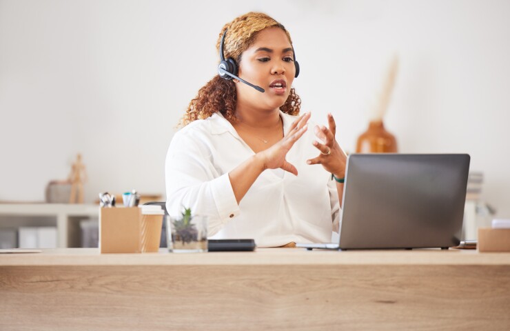 A woman sits at her wood desk, using her hands to gesture while speaking as she looks at her laptop. She has a headset on.