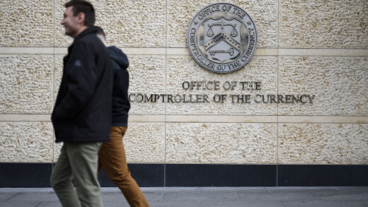 A picture of two people walking past signage outside the Office of the Comptroller of the Currency headquarters in Washington, DC.