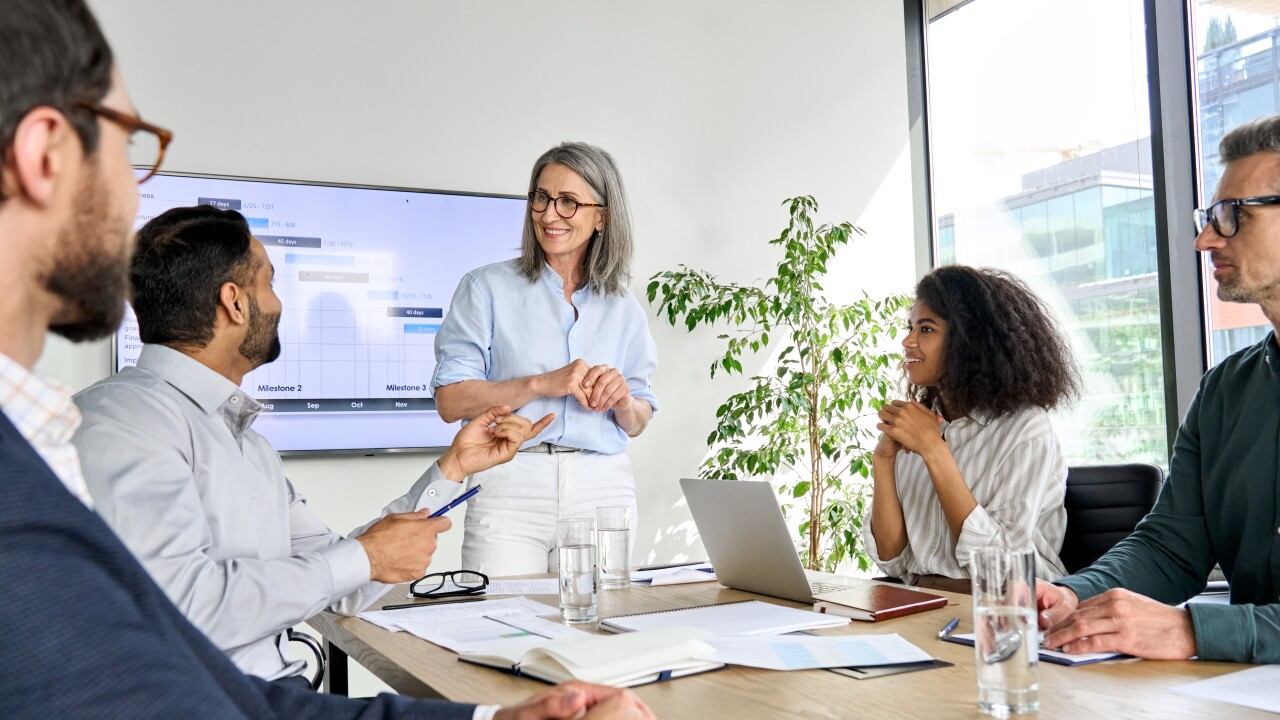 People in a meeting, sitting at a table with one person standing near a screen.