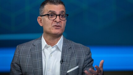 A man with glasses and a grey plaid blazer gestures while speaking against a blue, geometric patterned background.