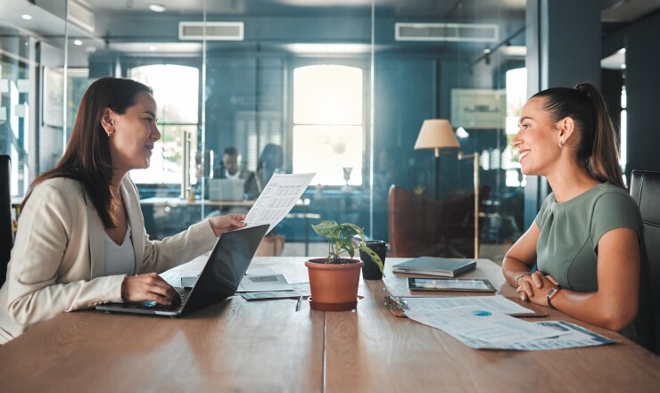 Businesswoman with laptop computer, holding paper and interacting with business colleague with papers on table