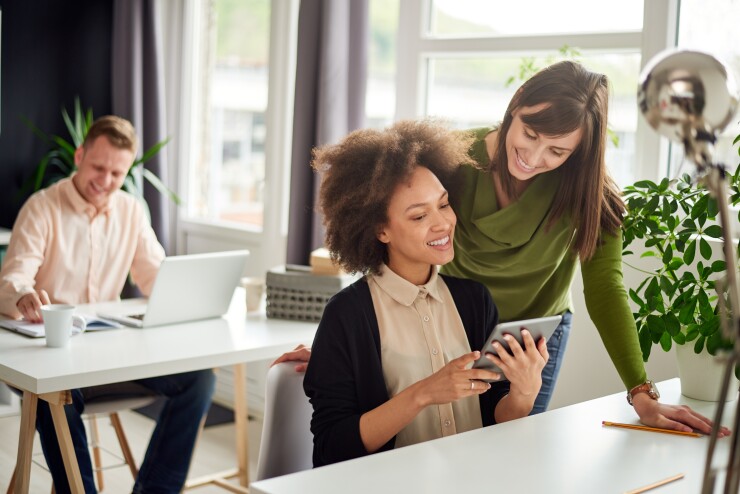 Two female employees looking at laptop man sitting at desk behin them