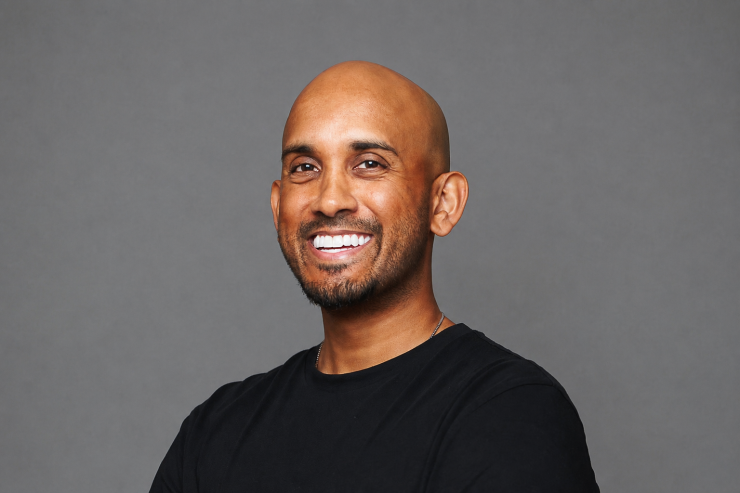 Smiling man with black shirt standing against a gray background.