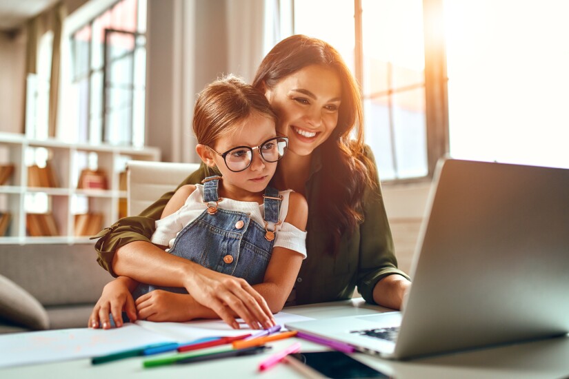 Mom holding daughter on lap, smiling and working on laptop computer