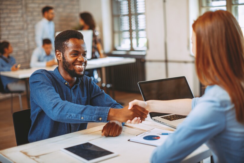 A Black man shakes the hand of the interviewer at a table.