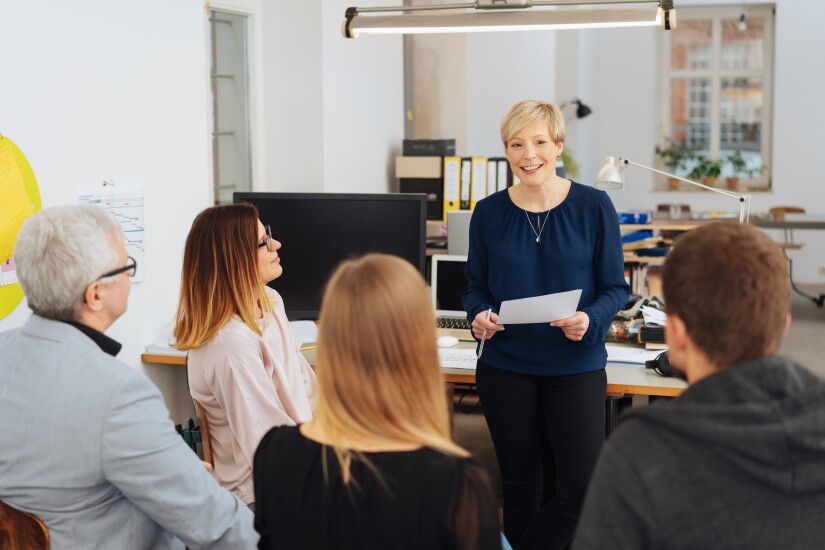 Group of seated employees listening to female employee who is smiling, leaning on desk