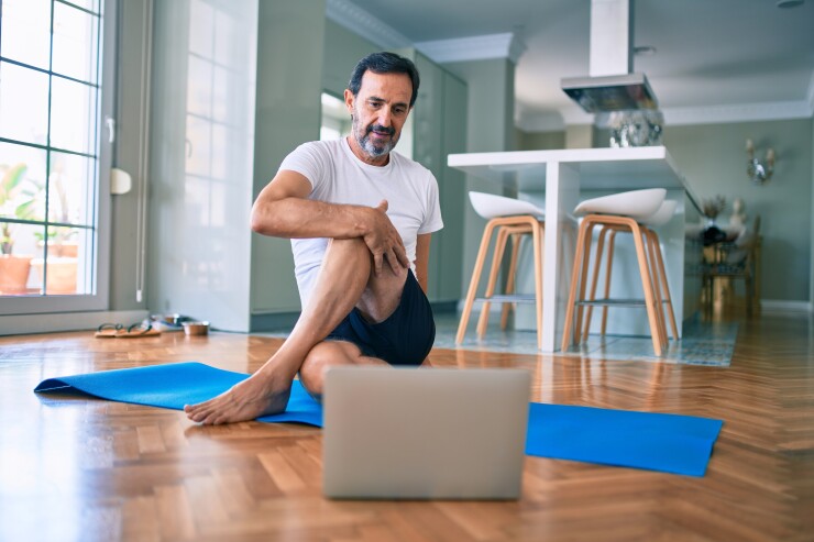 Man sitting on yoga mat stretching looking at laptop computer