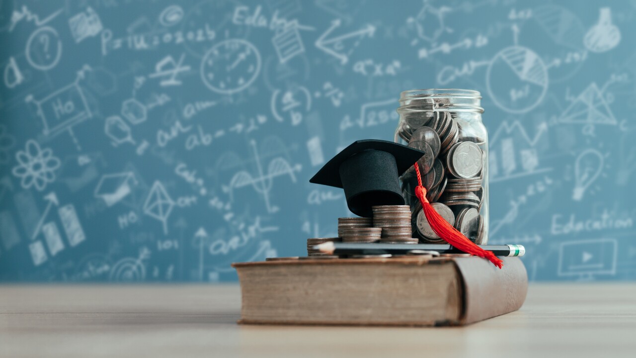 A book sits on a table with change and a graduate's hat, all in front of a chalkboard.