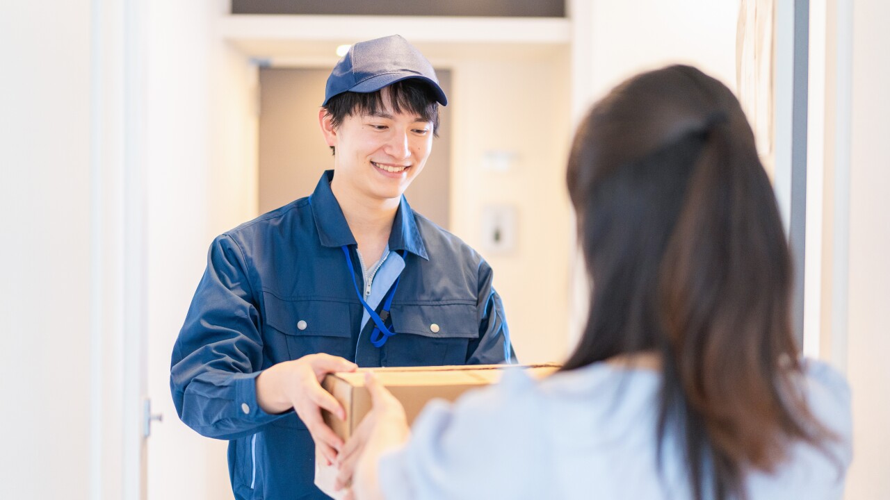 A smiling delivery man hands a brown-box package to a woman at the threshold of her apartment.