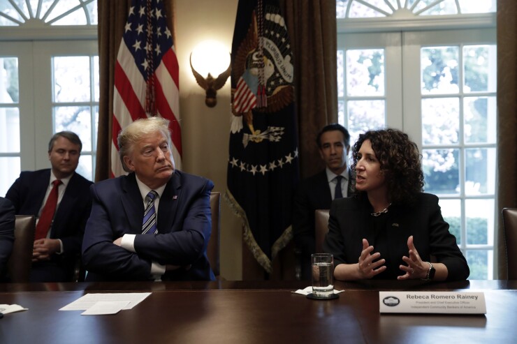Rebeca Romero Rainey sits at a table, speaking at an event with President Donald Trump.