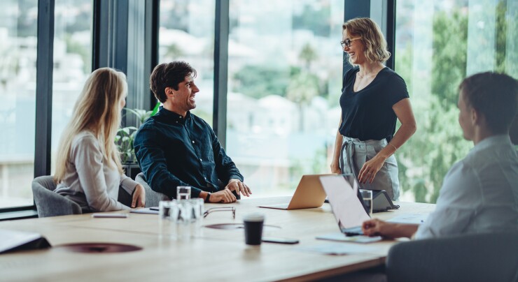 Group of employees sitting at table, woman standing, talking