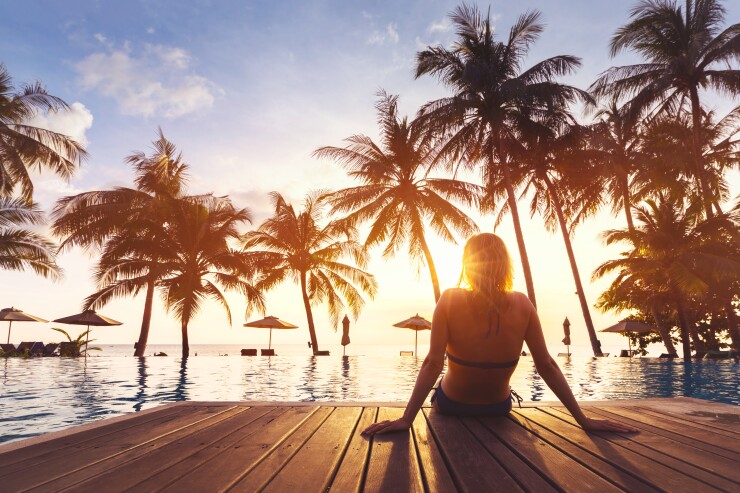 A woman in a bathing suit watches the sunset while sitting at the pool, just her legs in the water; there are palm trees in the distance.
