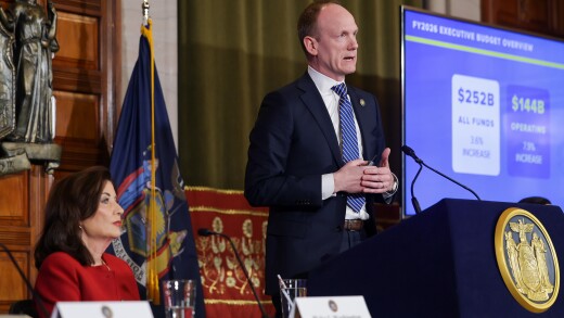 New York Gov. Kathy Hochul, seated, and budget director Blake Washington, standing at a podium.