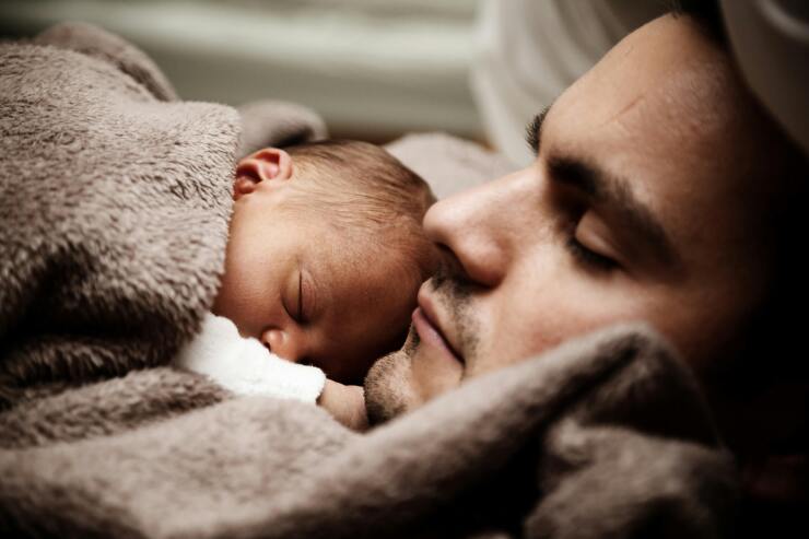 A father holds his newborn baby to his chest; they are both wrapped in a fluffy brown blanket.