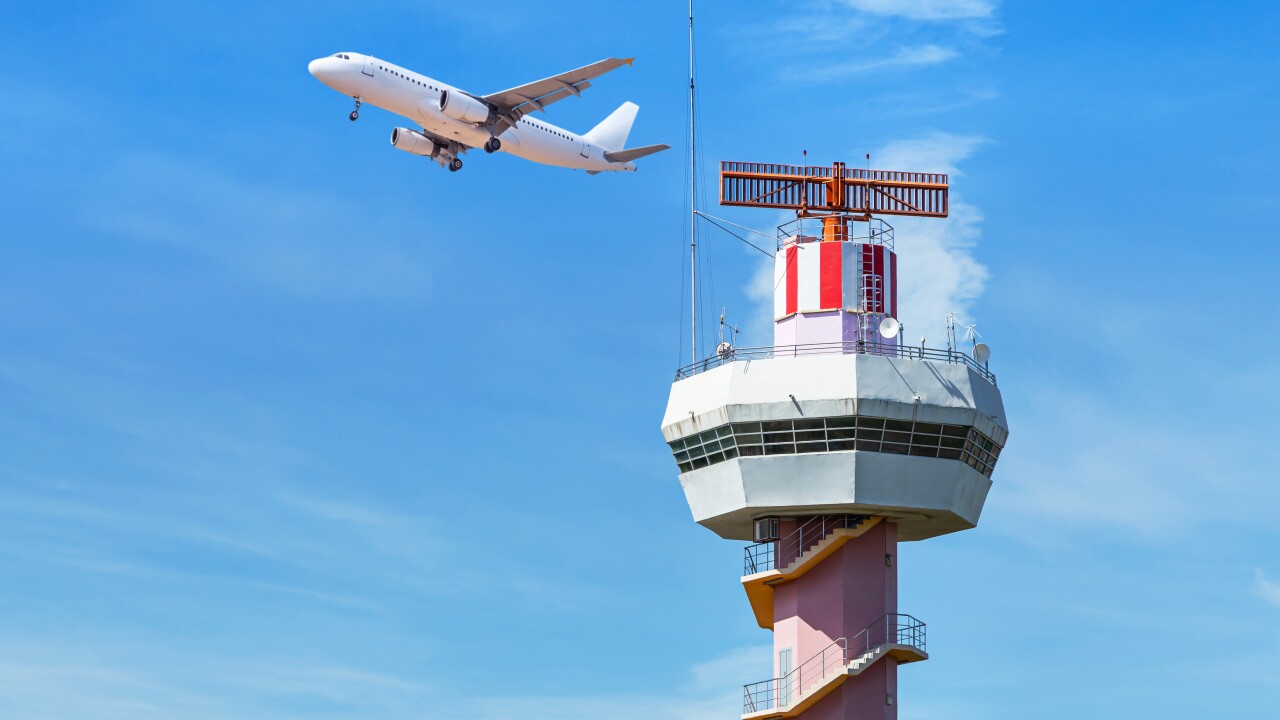 Airport traffic control tower in the foreground, a plane flies behind it, and the sky is blue.