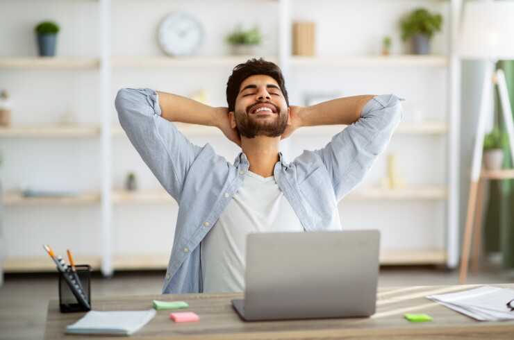 Man sitting at desk in front of computer leaning back with hands behind head smiling