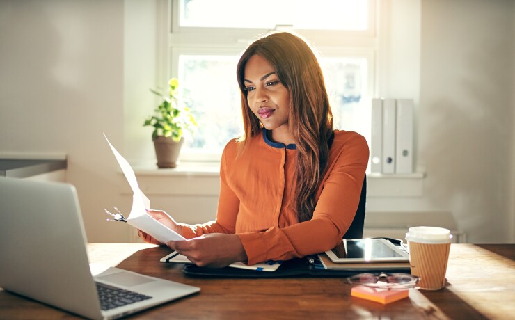 Woman sitting, working at desk, holding paper and looking at computer