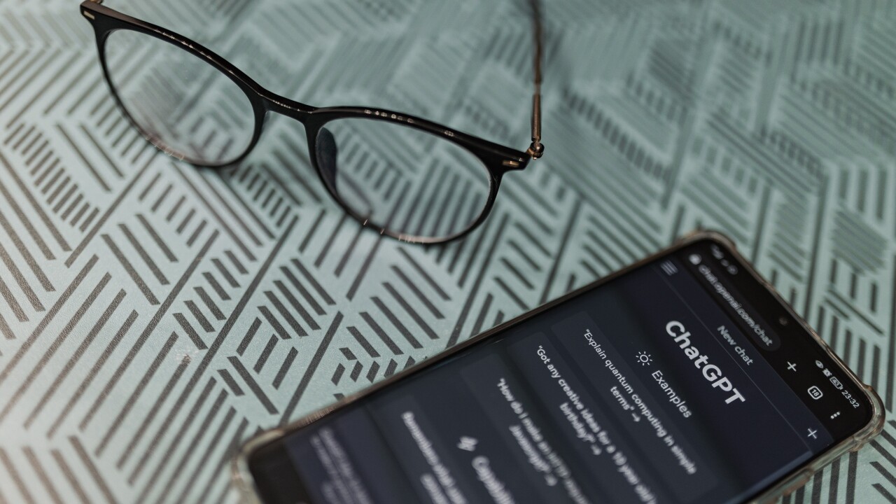 A pair of eyeglasses sits on a surface next to a smart phone that has ChatGPT pulled up on its screen.