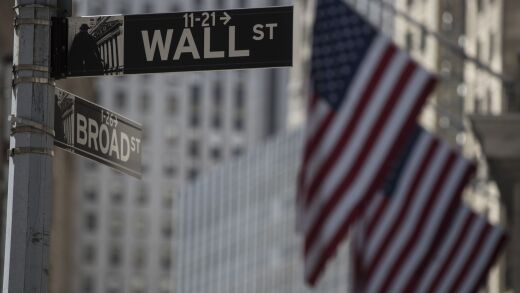 A Wall Street sign hangs in front of American flags outside the New York Stock Exchange (NYSE) in New York, U.S., on Monday, July 31, 2017. U.S. stocks fluctuated to end a month of gains spurred by corporate results and growing optimism in the strength of the global economy. Photographer: Victor J. Blue/Bloomberg