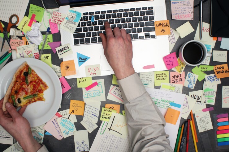 A person eats pizza while working at a laptop surrounded by to-do notes.