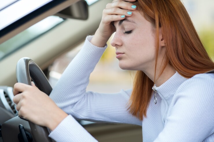 A distressed teenager has one hand on the steering wheel of a car and another hand on her forehead in frustration.