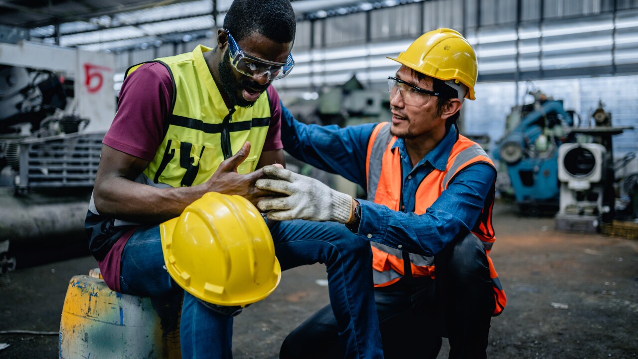 Two construction workers in yellow hard hats; one worker injured his finger, and the other is kneeling next to him.