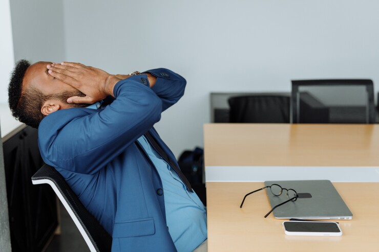 A man sits at a desk with his laptop closed. He is leaning back in stress and rubbing his face with his hands.