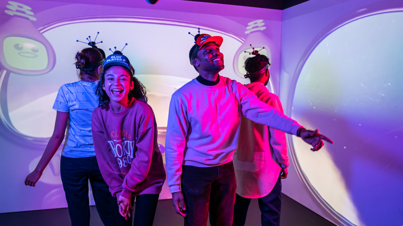 A group of four friends with visors on their head are looking around a digital game room -- a room filled with screens with cartoon characters on it.