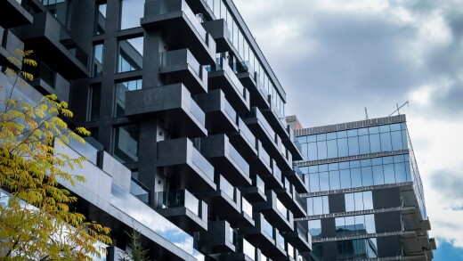 Modern, dark gray facade of a high-rise residential building with projecting balconies and large glass windows, set against a dramatic, cloudy sky, with a touch of autumn foliage.