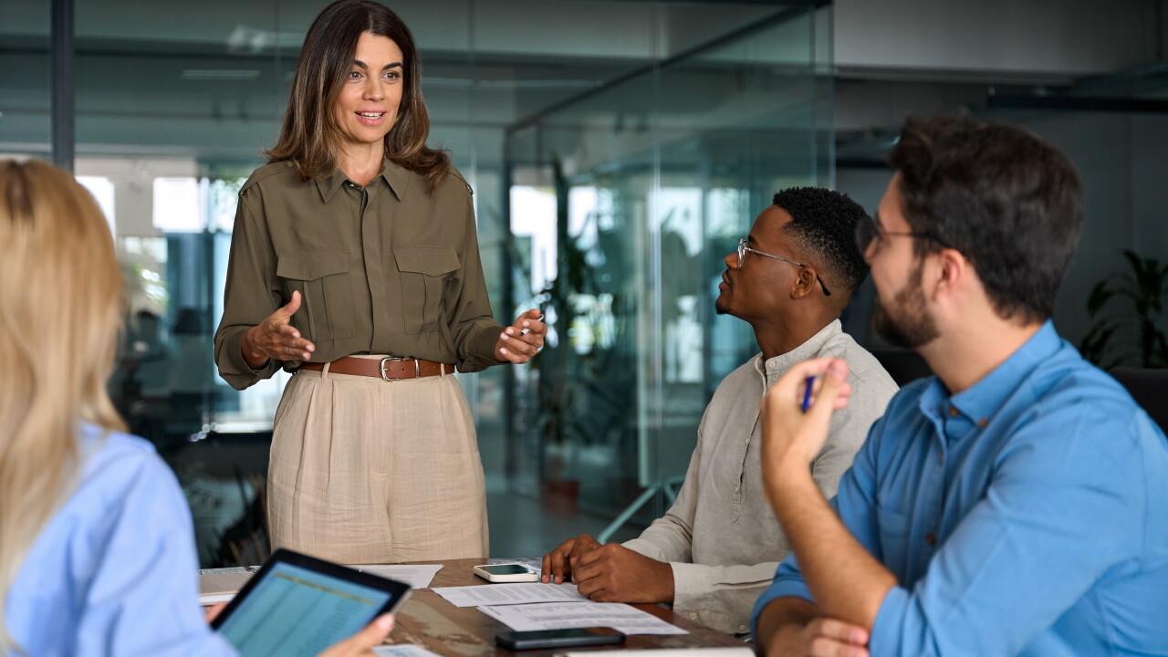 Group of employees sitting around table, woman standing talking