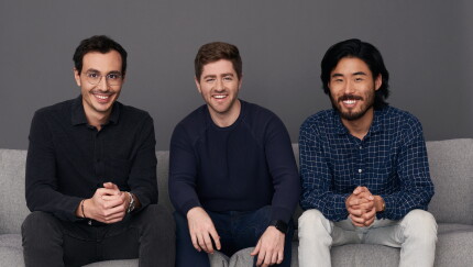 Three men smile while sitting side by side on a grey sofa against a plain dark grey background.