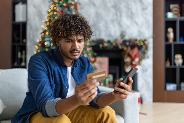 Cheated and upset Christmas man in living room near decorated Christmas tree, rejected and wrong money transfer, hispanic man holding bank credit card and phone