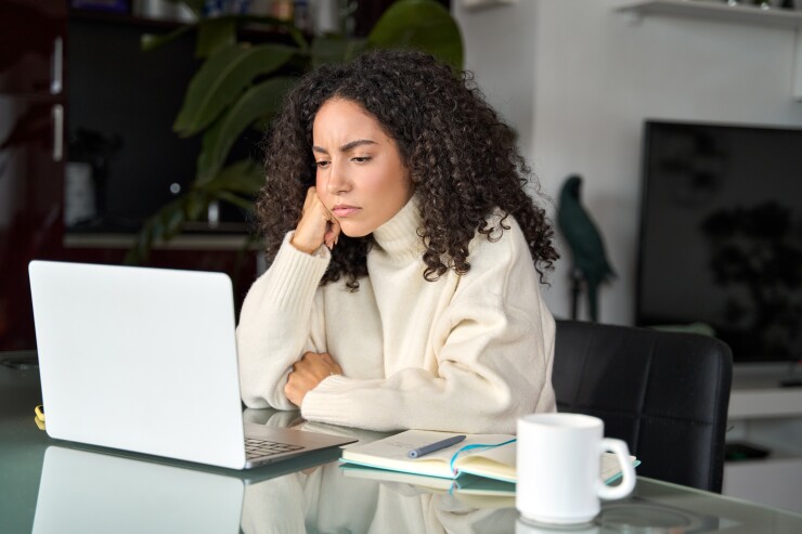 A woman in a white turtle neck rests her cheek on her hand while looking down at her laptop in frustration.